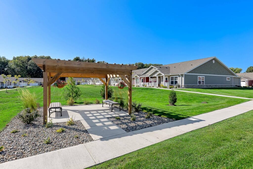 A wooden pergola with benches sits on a stone-covered area beside a sidewalk, with grass, shrubs, and a single-story building in the background under a clear blue sky.