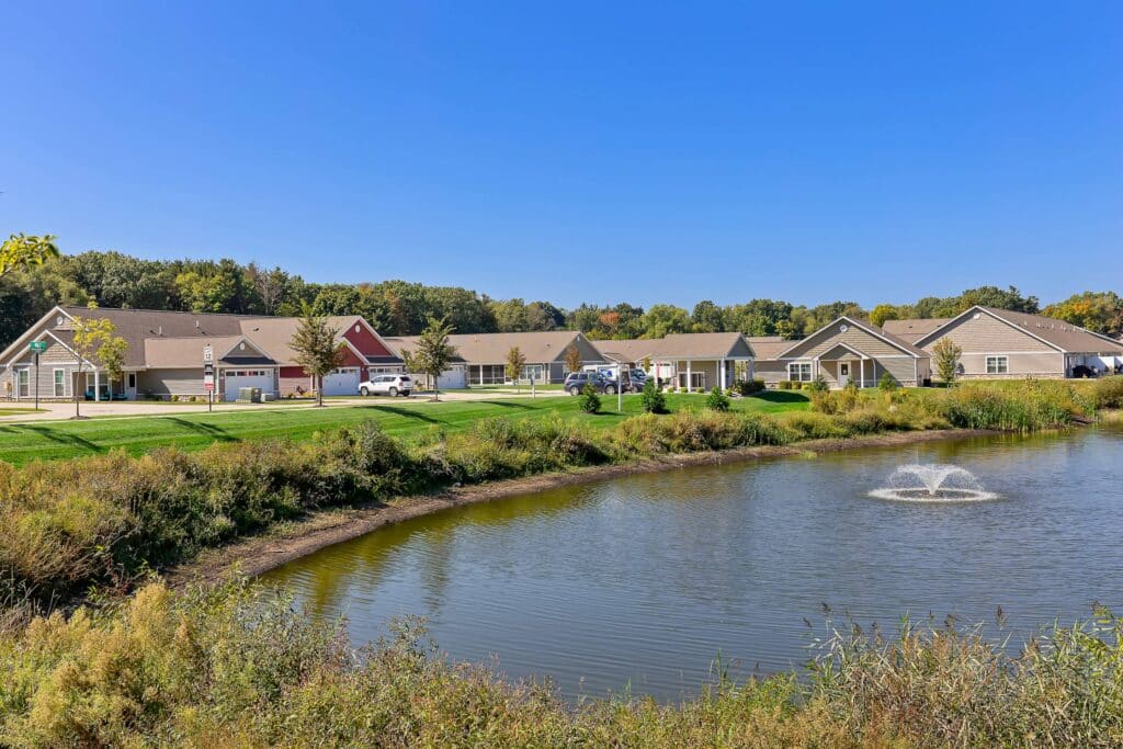 Row of single-story houses with garages facing a small pond and fountain, surrounded by greenery under a clear blue sky.