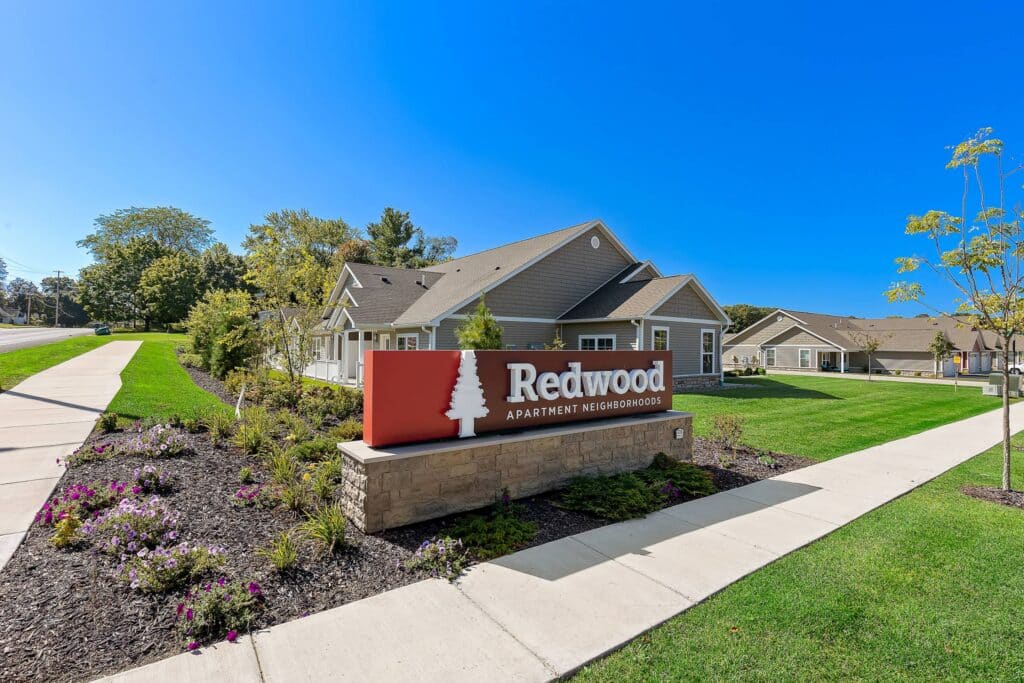 A sign reading "Redwood Apartment Neighborhoods" stands in front of a one-story residential building with manicured lawns and flower beds under a clear blue sky.