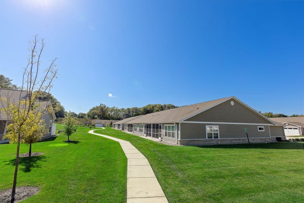 Sidewalk runs through a grassy area beside single-story tan residential buildings under a clear blue sky.