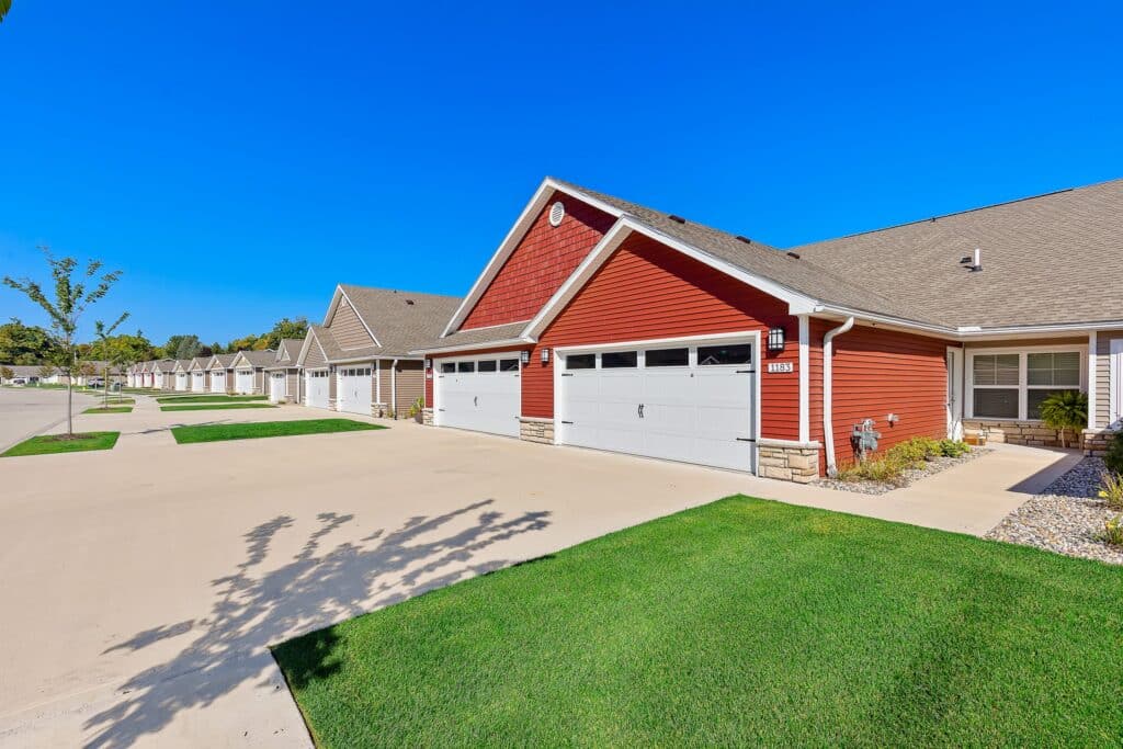 Row of modern, single-story red townhouses with attached garages, neatly trimmed lawns, and a clear blue sky.