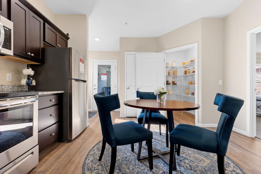 A modern kitchen with dark cabinets, stainless steel appliances, a round dining table with four blue chairs, and an open pantry with organized shelves.