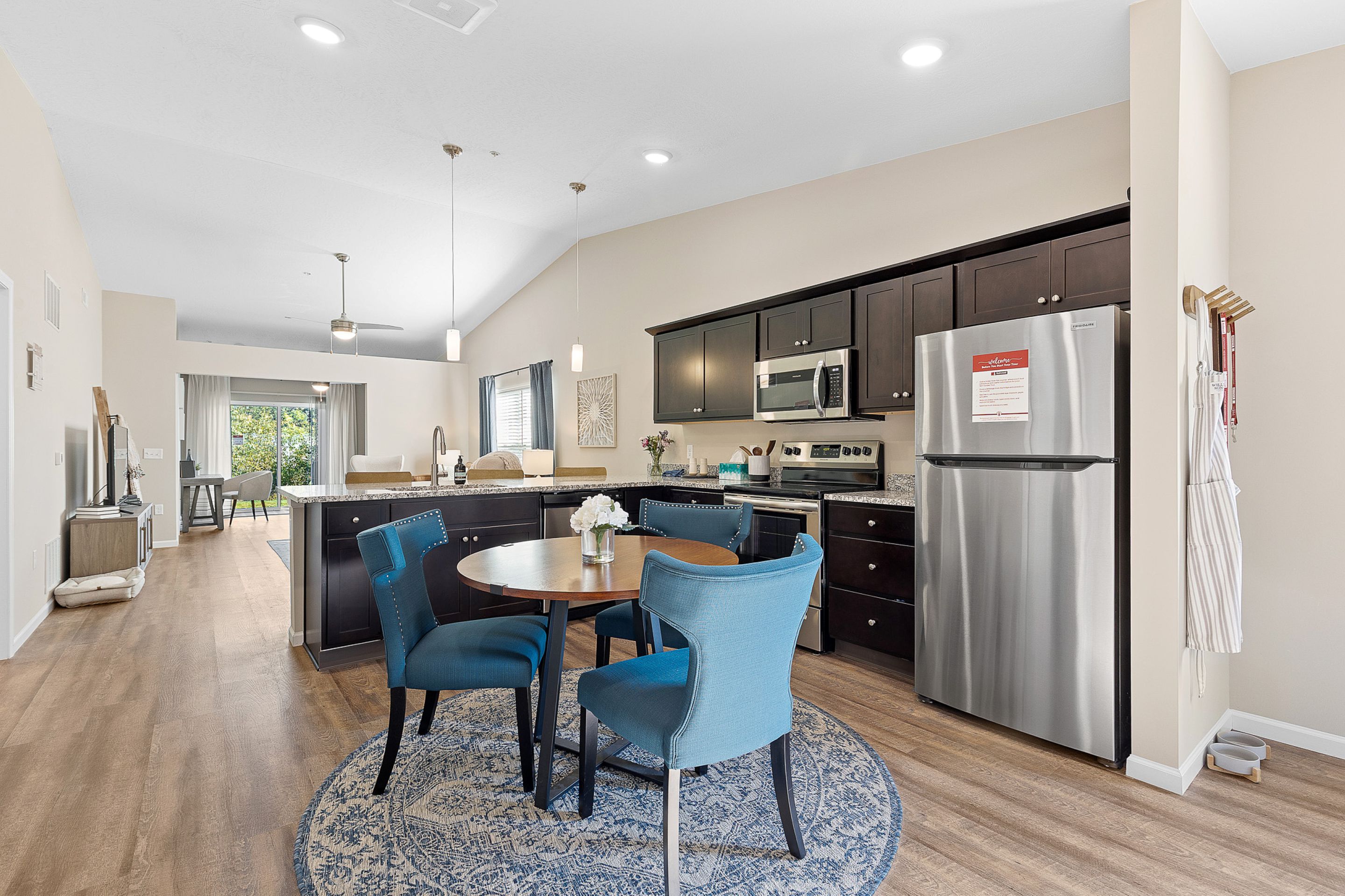 Modern kitchen and dining area with stainless steel appliances, dark cabinets, round table with blue chairs, and wood flooring, open to a living space with large windows.