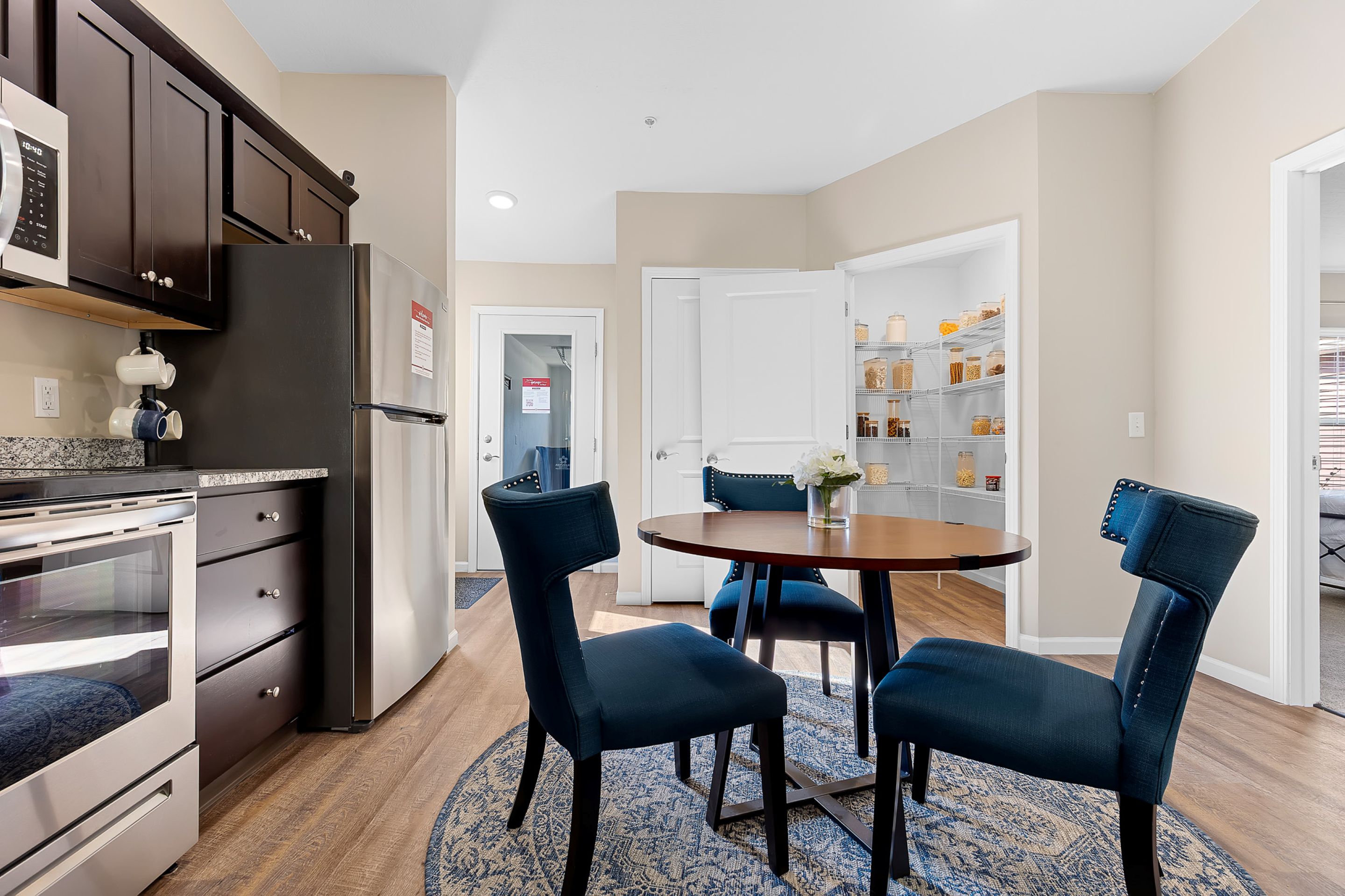 A modern kitchen with dark cabinets, stainless steel appliances, a round dining table with four blue chairs, and an open pantry with organized shelves.