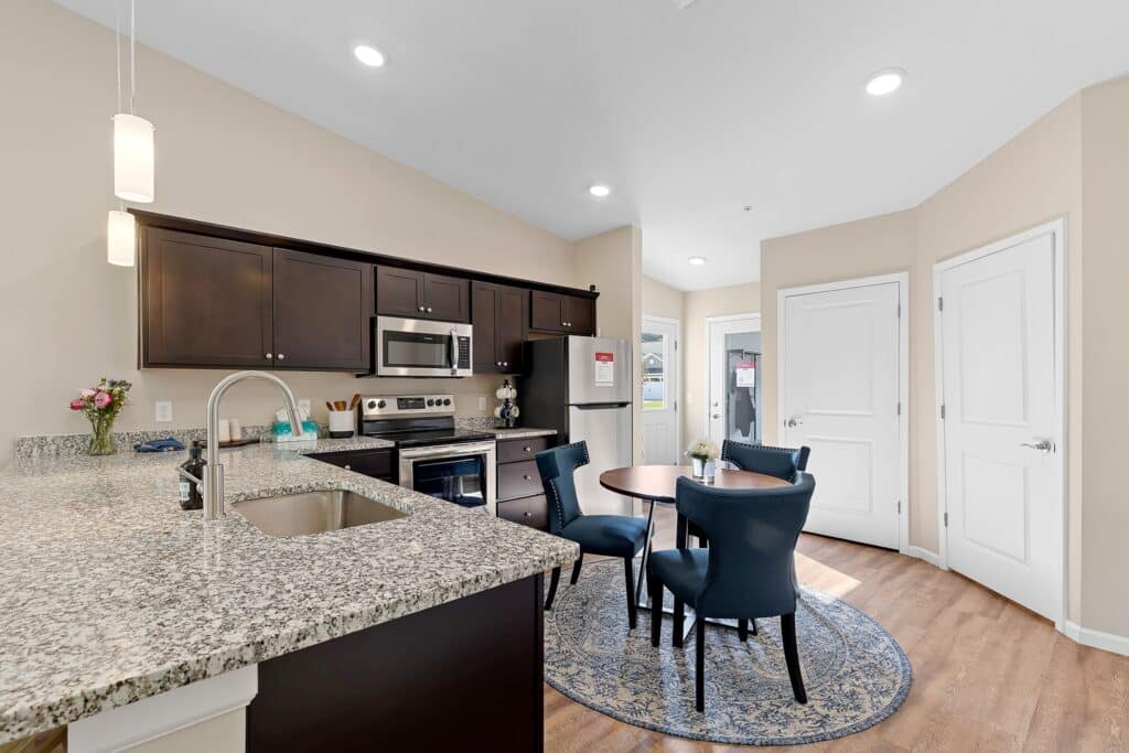 Modern kitchen with granite countertops, stainless steel appliances, dark wood cabinets, a round dining table with four blue chairs, and wood flooring.