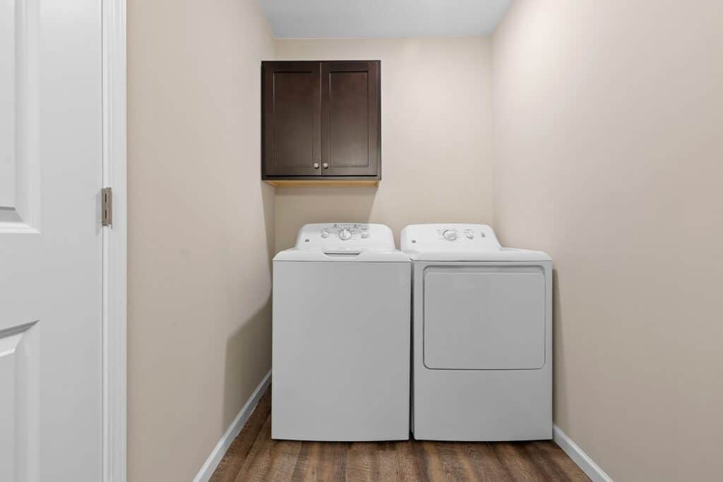 A laundry room with a white washing machine and dryer side by side, a dark wood cabinet above them, and light beige walls with wood flooring.