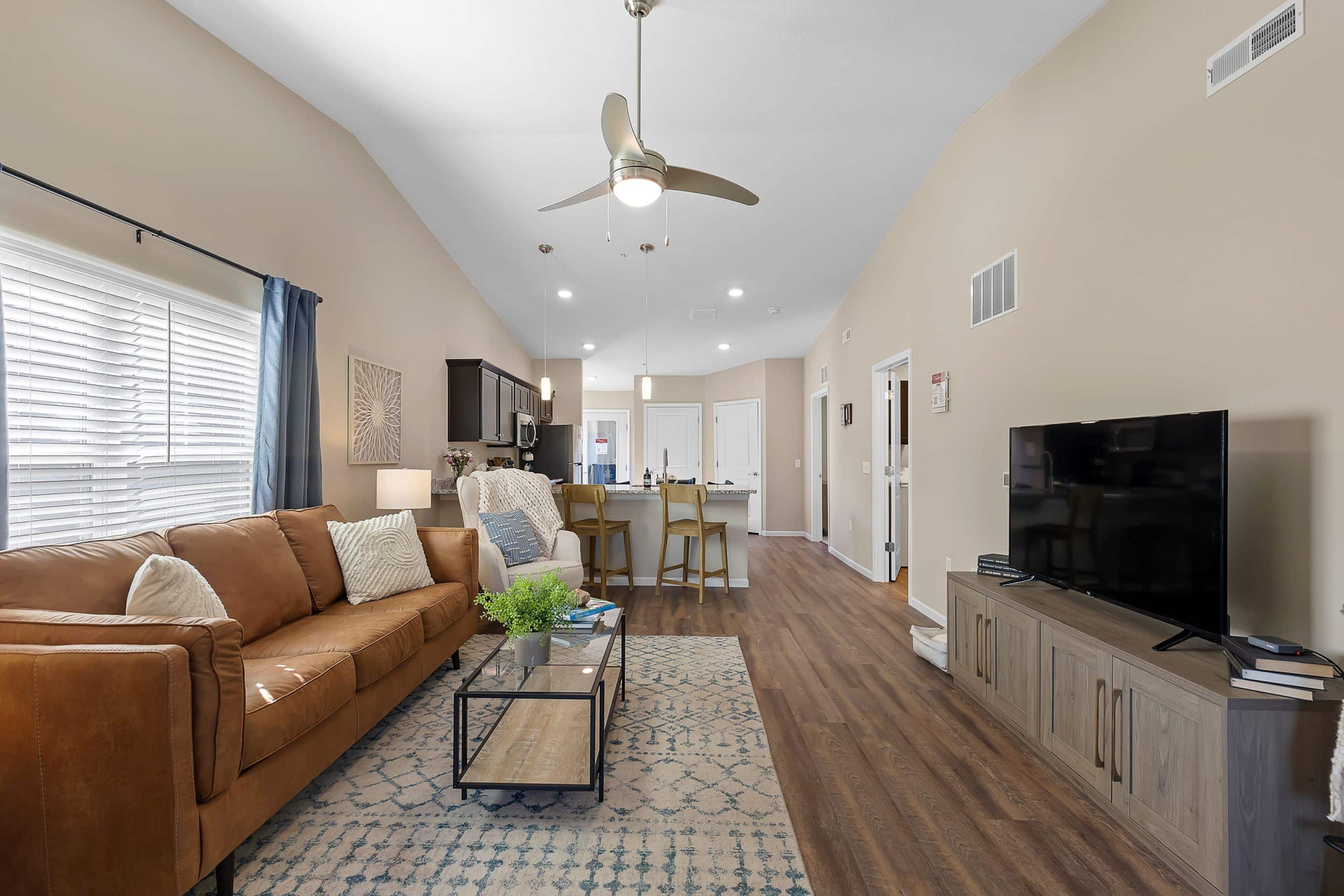 Modern living room with tan walls, brown leather sofa, wall-mounted TV, glass coffee table, and open kitchen with bar stools in the background. Wood flooring throughout the space.