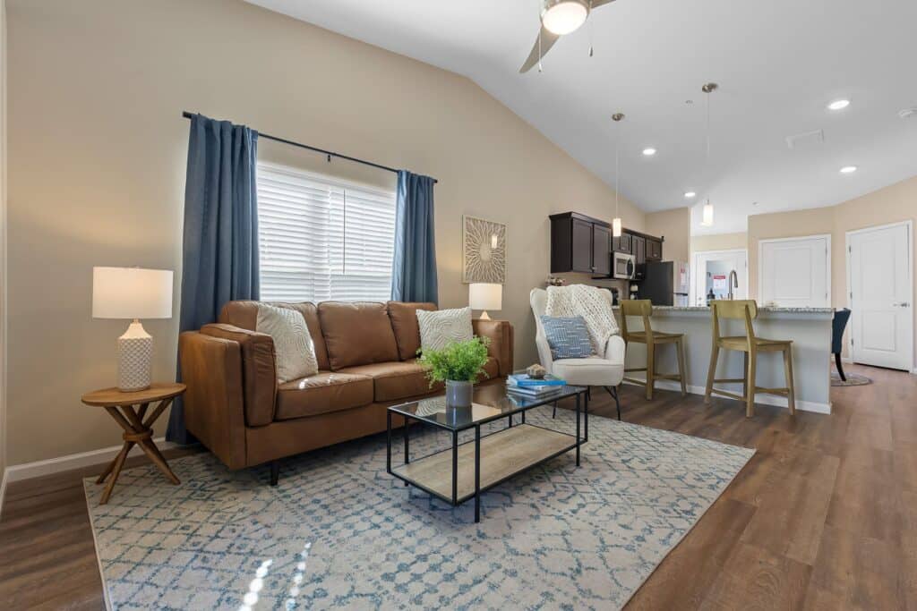 Modern living room with brown sofa, glass coffee table, rug, and open kitchen area featuring barstools and pendant lighting. Wood flooring throughout, with neutral tones and blue accents.