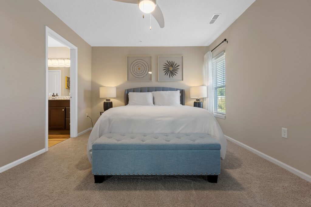 A neatly made bed with white bedding, a blue tufted bench at the foot, two matching lamps on nightstands, and two framed artworks above the headboard in a beige bedroom.
