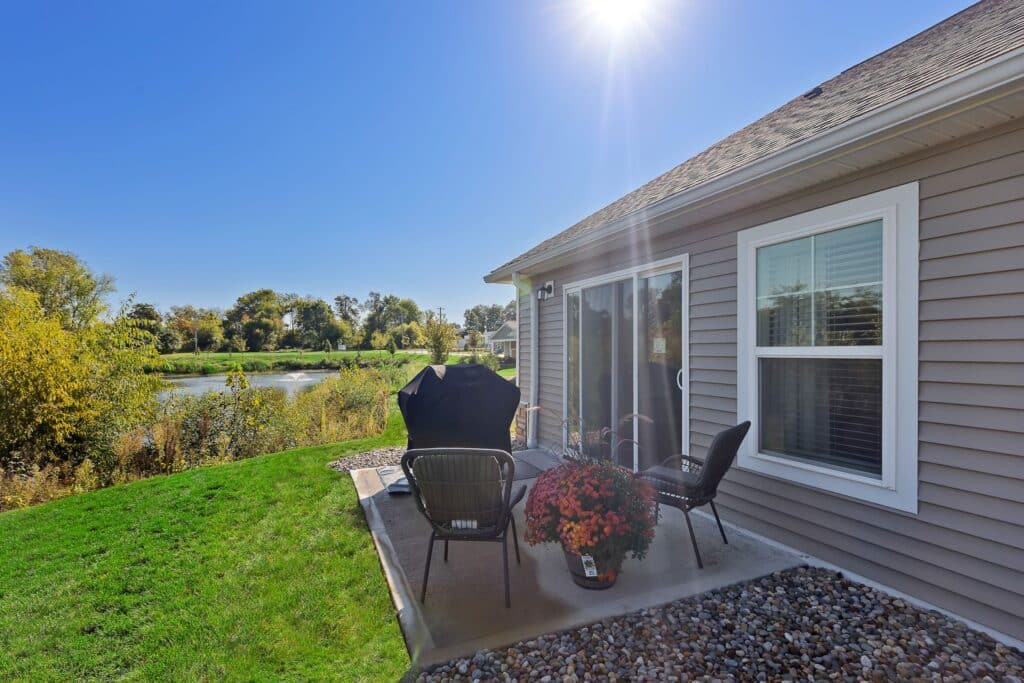 Small backyard patio with four chairs, a grill, a potted plant, and sliding glass door, overlooking grass, trees, and a pond under a clear sunny sky.
