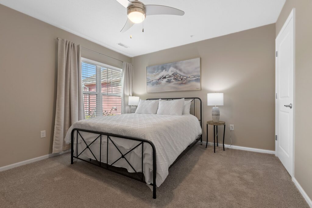 A bedroom with a metal framed bed, white bedding, two bedside tables with lamps, beige walls, a window with curtains, and a ceiling fan.