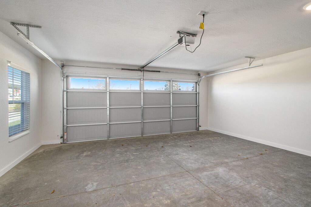 Empty two-car garage with a closed metal door, white walls, concrete floor, two ceiling-mounted openers, and a window on the left wall letting in natural light.