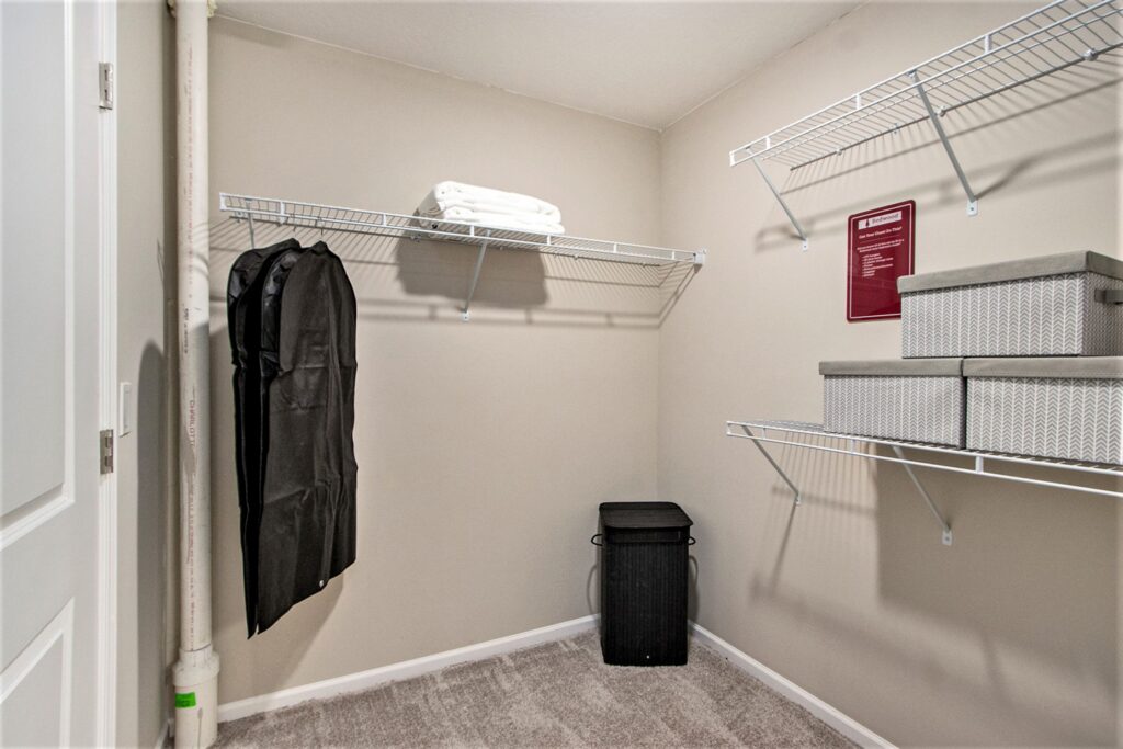 A walk-in closet with wire shelves, a garment bag, folded white towels, three gray storage boxes, a small black laundry hamper, and a beige carpeted floor.