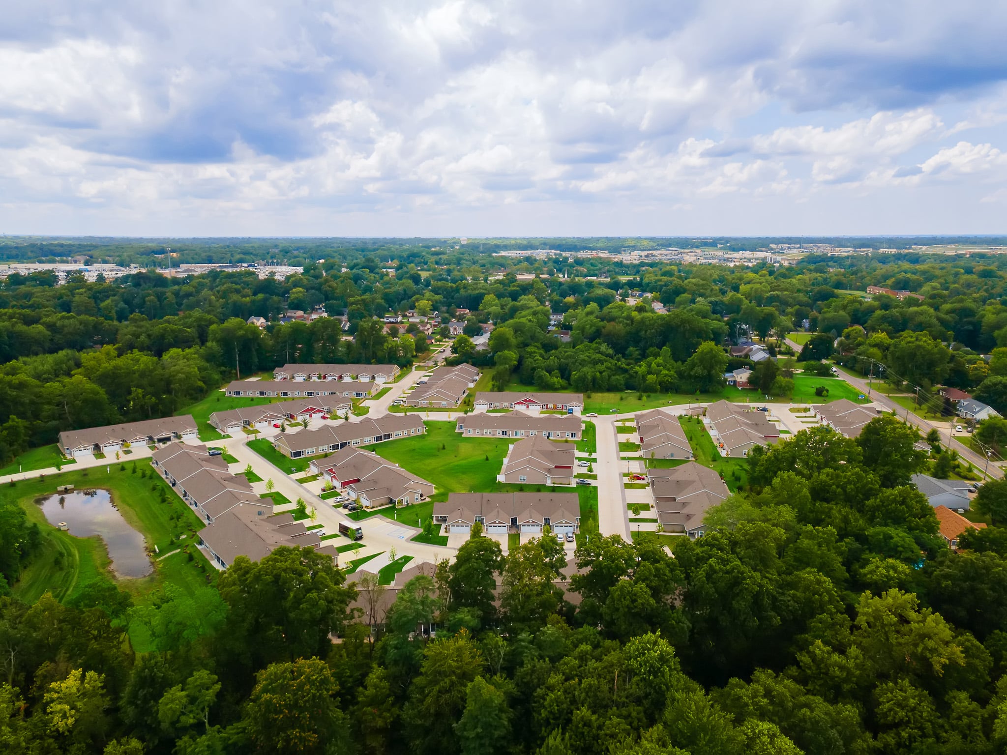 Aerial view of a suburban neighborhood with rows of houses, green lawns, trees, and a small pond, under a partly cloudy sky.