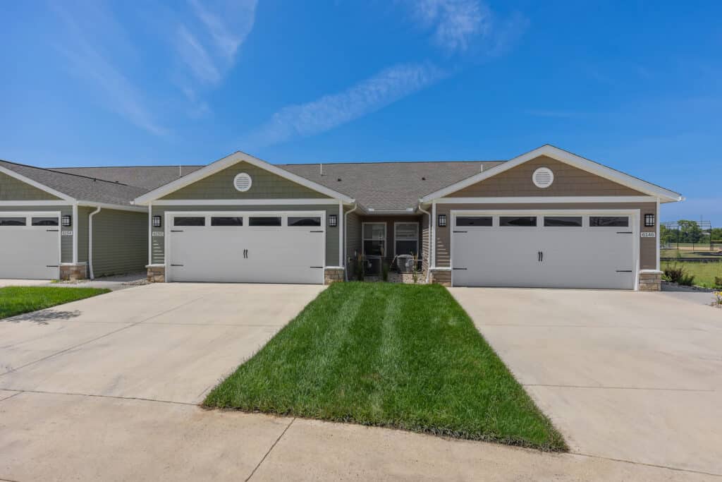 Duplex with two attached garages, green lawns, concrete driveways, and a clear blue sky in the background.