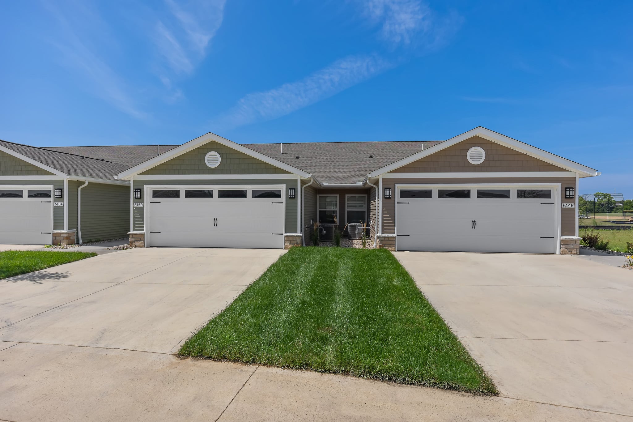 Duplex with two attached garages, green lawns, concrete driveways, and a clear blue sky in the background.