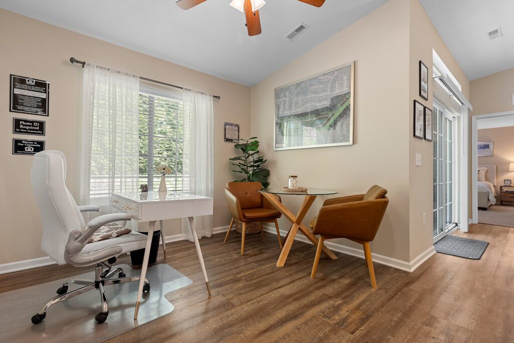 A home office with a white desk and chair, two brown chairs with a round table, indoor plant, wall art, wood flooring, and large window with sheer curtains.