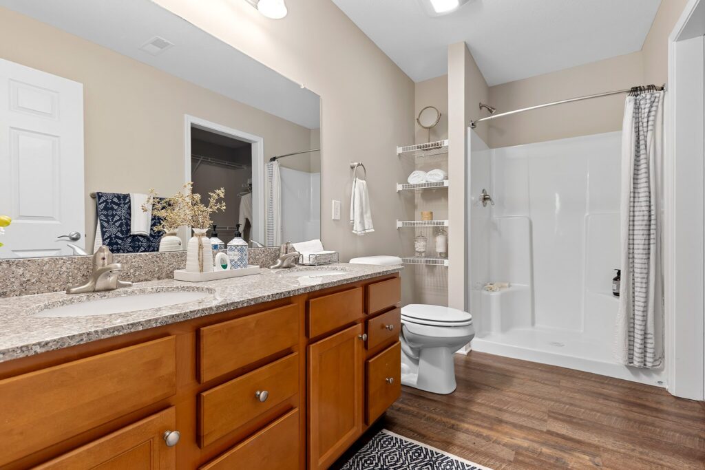Modern bathroom with double sinks, wooden cabinets, large mirror, toilet, walk-in shower with curtain, and shelves with towels on a wood-look floor.