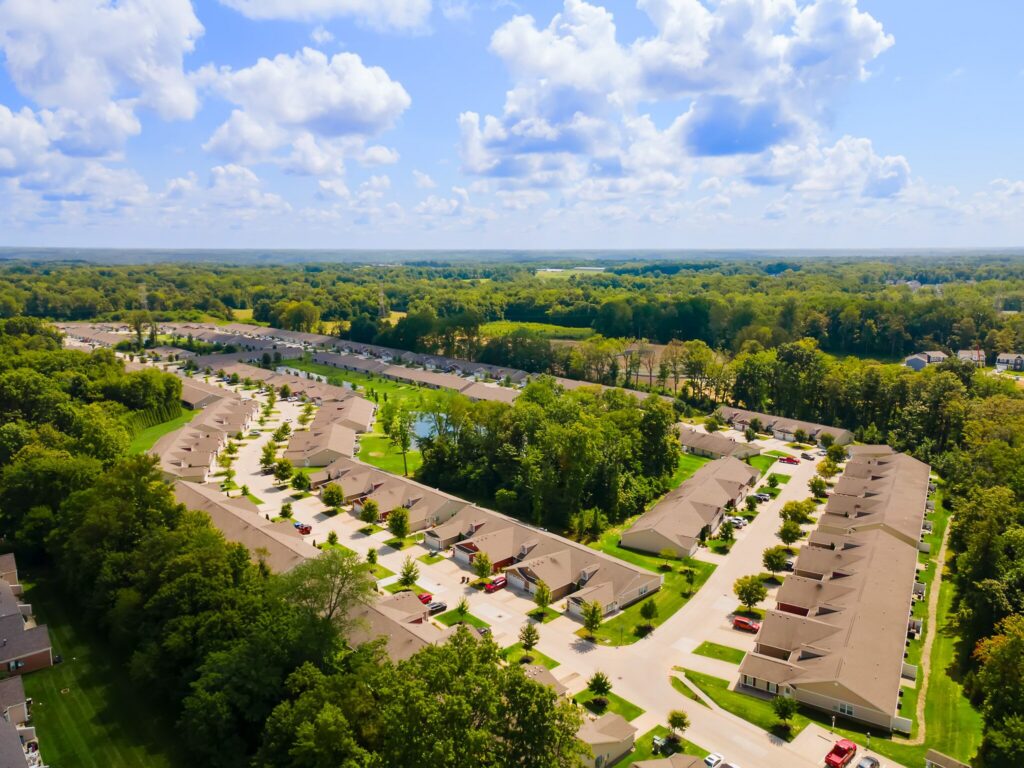 Aerial view of a suburban neighborhood with rows of houses, tree-lined streets, and patches of greenery under a partly cloudy sky.