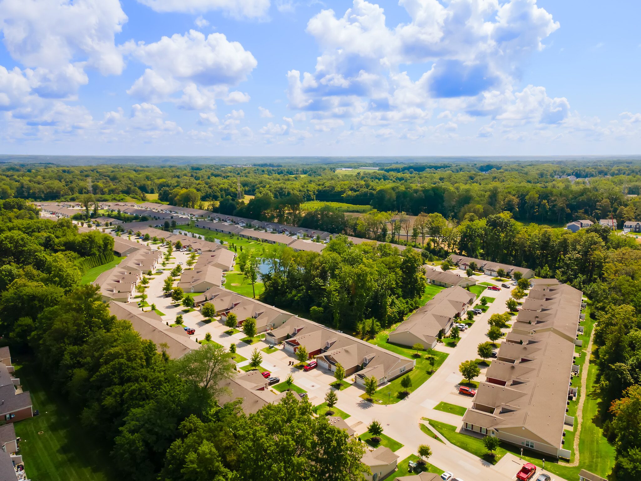 Aerial view of a suburban neighborhood with rows of houses, tree-lined streets, and patches of greenery under a partly cloudy sky.
