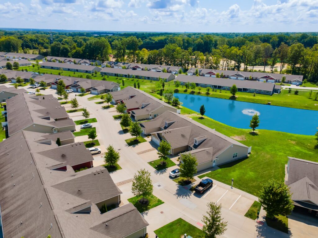 Aerial view of a residential neighborhood with rows of houses, a pond with a fountain, green lawns, and parked cars on sunny day.
