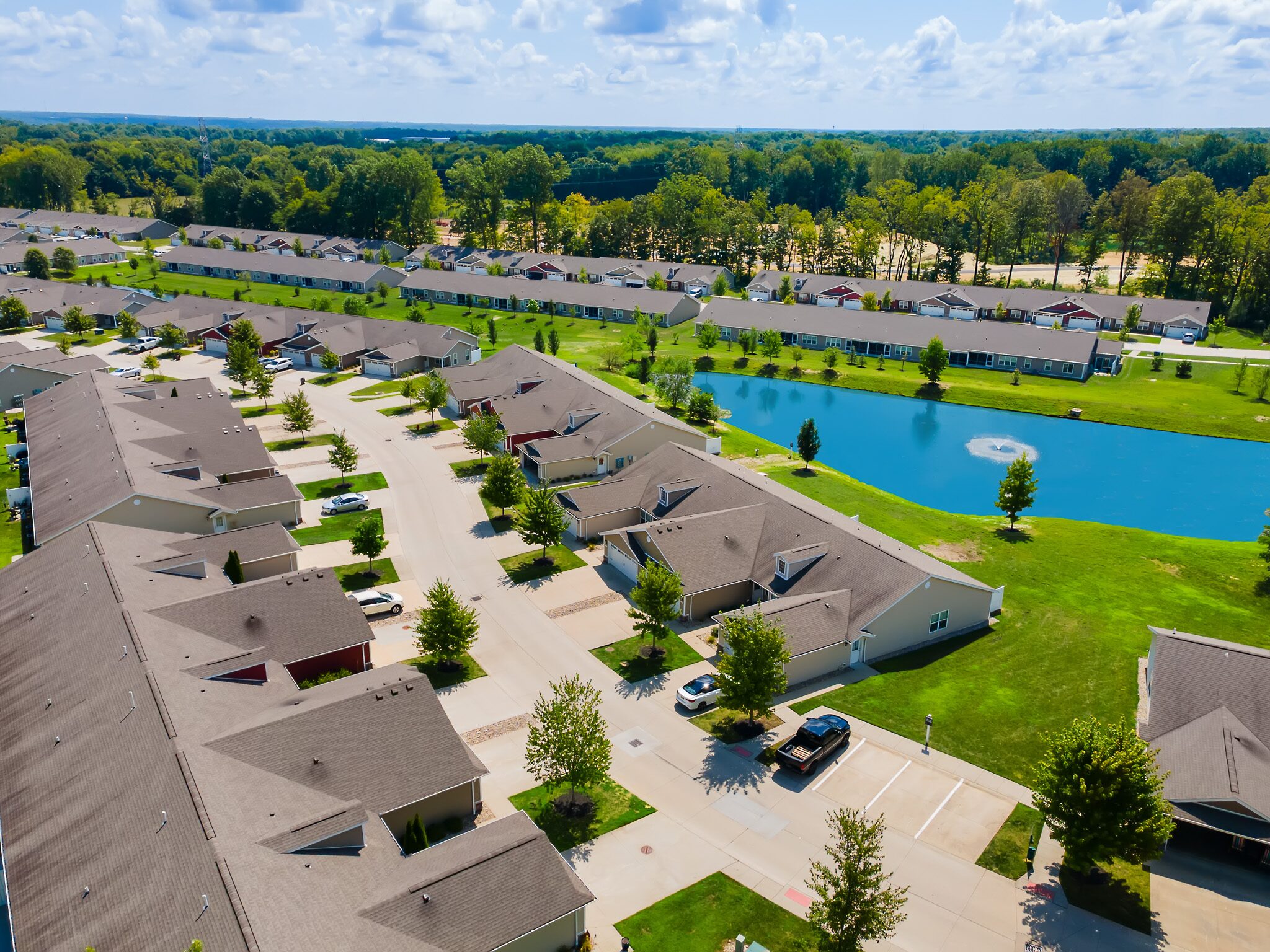 Aerial view of a residential neighborhood with rows of houses, a pond with a fountain, green lawns, and parked cars on sunny day.
