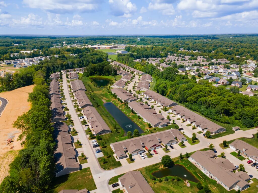 Aerial view of a suburban neighborhood with rows of houses, green lawns, small ponds, and surrounding trees under a partly cloudy sky.