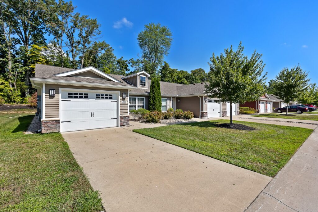 Single-story house with attached garage, concrete driveway, manicured lawn, small tree, and clear blue sky. Neighboring homes visible in the background.