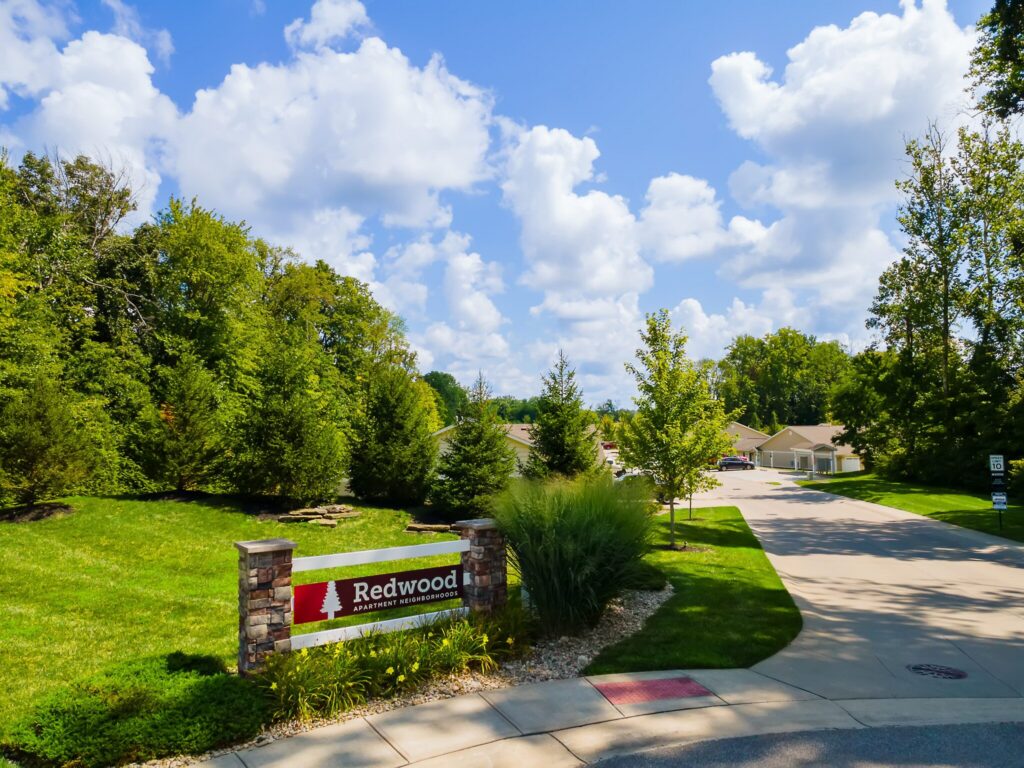 Entrance to Redwood apartment neighborhood with sign, green lawn, trees, and a paved driveway leading to residential buildings under a blue sky with scattered clouds.