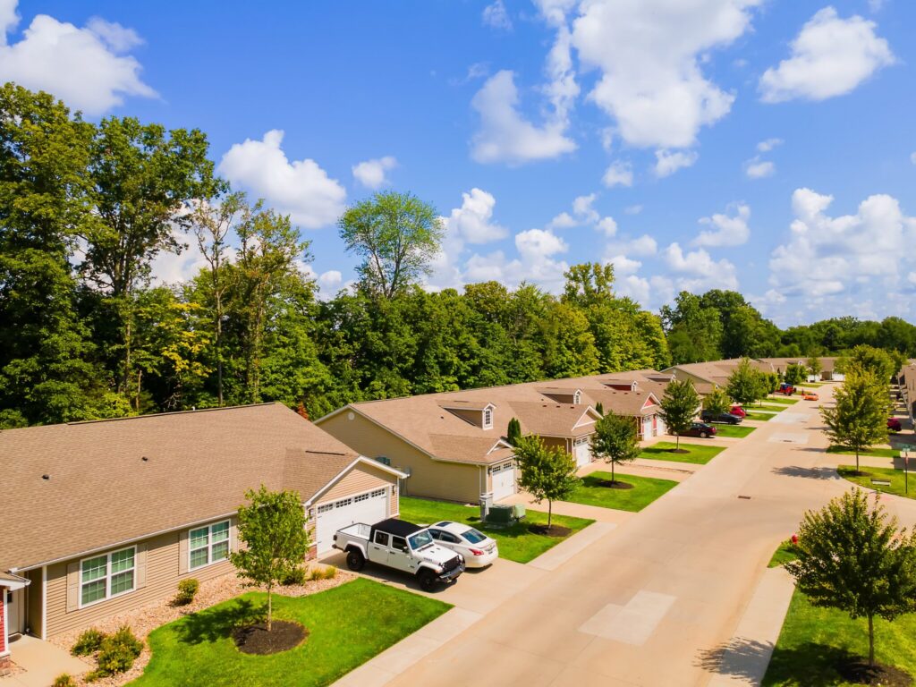A suburban neighborhood with single-story houses, driveways with parked cars, green lawns, and a tree line in the background under a partly cloudy sky.