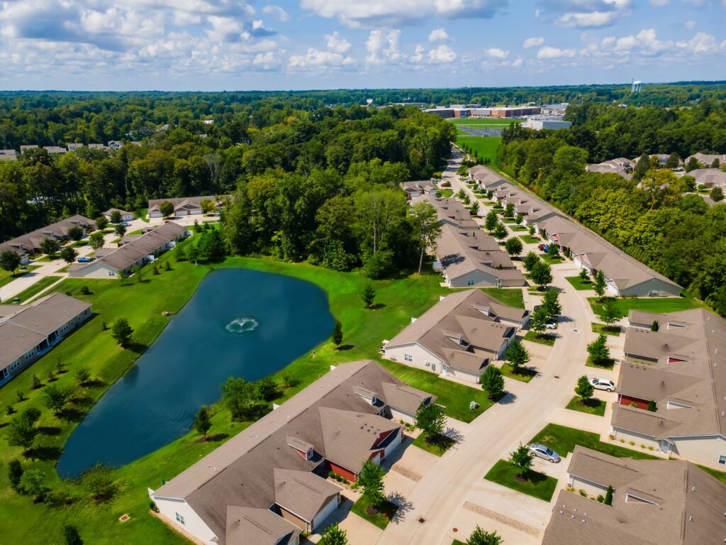 Aerial view of a suburban neighborhood with rows of houses, a small pond with a fountain, green lawns, trees, and a school building in the background under a partly cloudy sky.