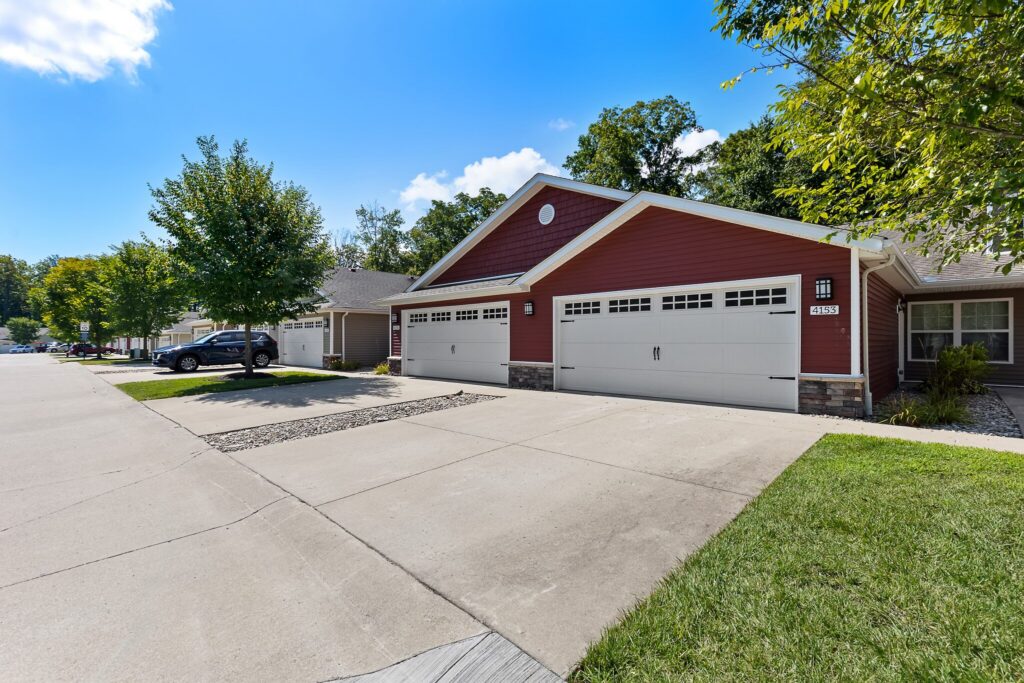 A red single-story house with three white garage doors, a wide concrete driveway, and green lawn under a blue sky with scattered clouds.