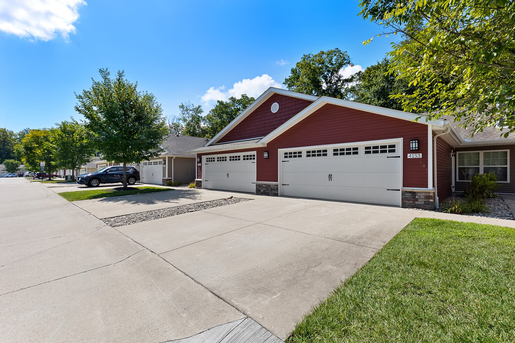 A red single-story house with three white garage doors, a wide concrete driveway, and green lawn under a blue sky with scattered clouds.