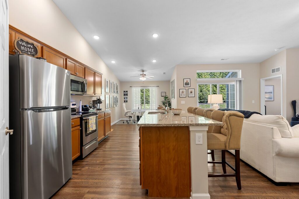 Modern kitchen with stainless steel appliances, wooden cabinets, granite island with bar stools, and an open living area featuring a sofa and large windows with greenery outside.