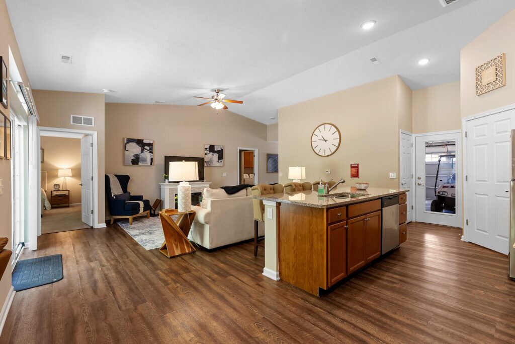 Open-concept living room and kitchen with wood flooring, beige walls, ceiling fan, and modern furnishings. A large clock hangs on the wall above the kitchen island.