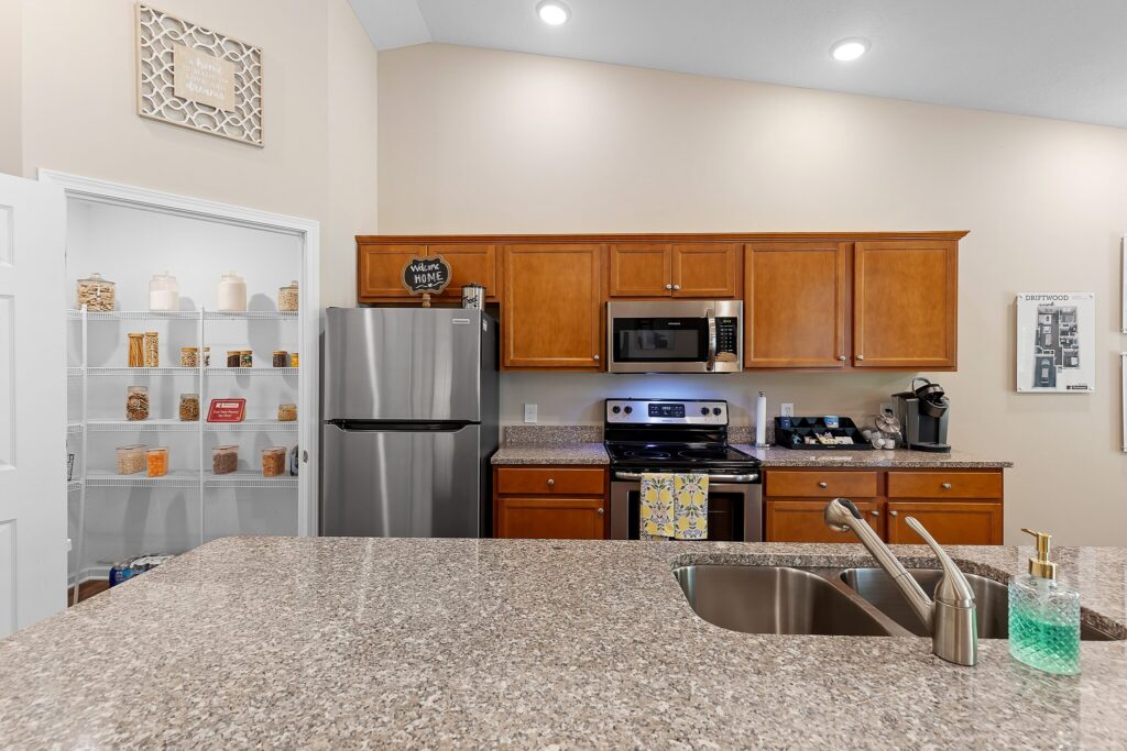 Modern kitchen with brown cabinets, stainless steel appliances, granite countertops, and a double sink; a pantry with organized containers is visible through an open door.