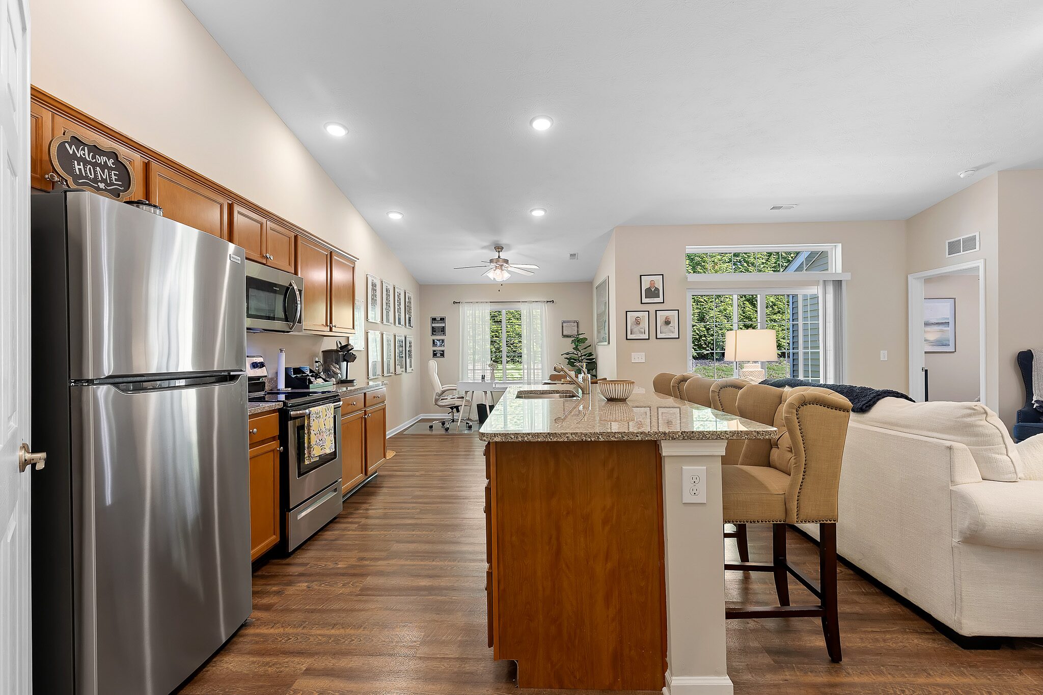 Modern kitchen with stainless steel appliances, wooden cabinets, granite island with bar stools, and an open living area featuring a sofa and large windows with greenery outside.