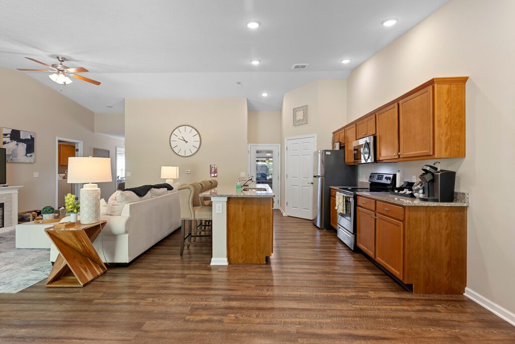 Open-concept kitchen and living room with wood floors, brown cabinets, stainless steel appliances, a beige sectional sofa, and neutral-colored walls.