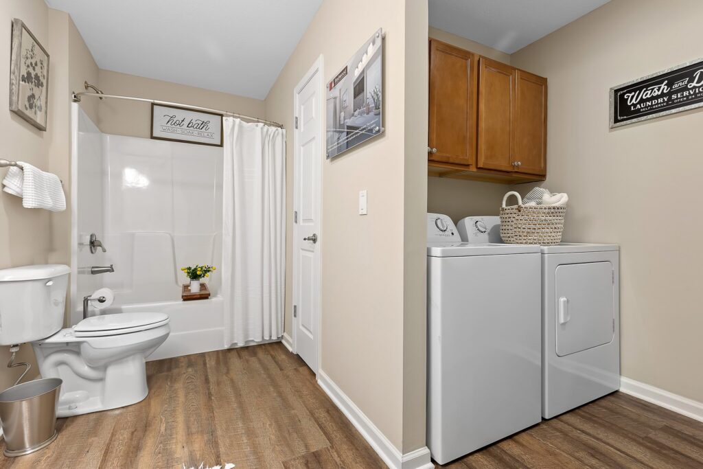 Bathroom and laundry room combination with a toilet, shower, washer, dryer, wood cabinets, and wall decor; the floor is wood-look vinyl and walls are beige.