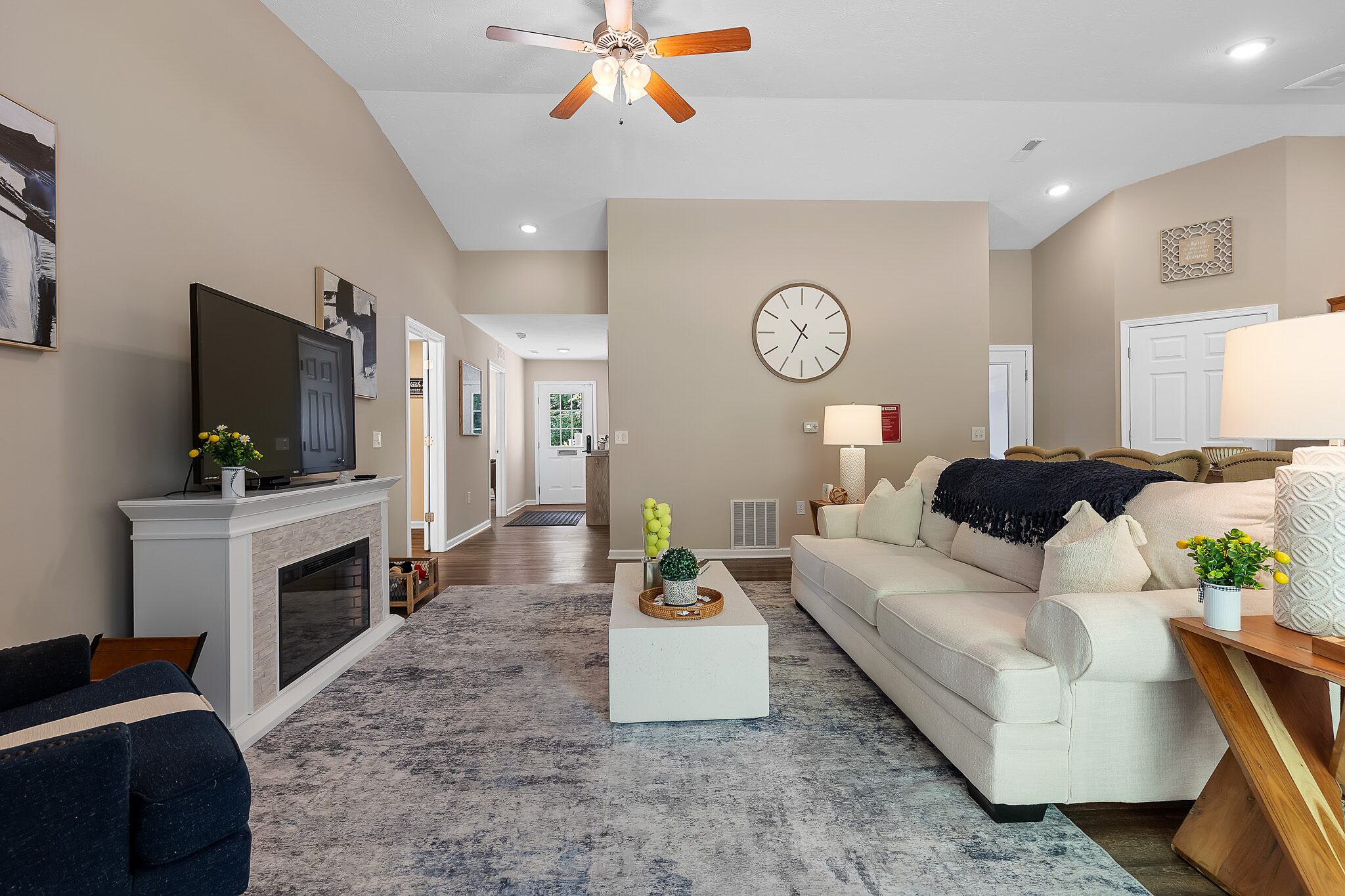 Modern living room with beige walls, white sofa, wall clock, TV on a fireplace, coffee table, and decorative accents. Ceiling fan and hallway visible in the background.