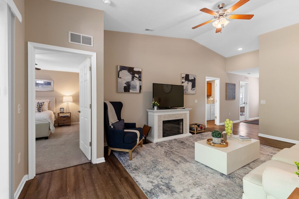 A modern living room with a TV on a white console, abstract wall art, an armchair, a rug, and a view into a bedroom and hallway. Ceiling fan with lights is on.