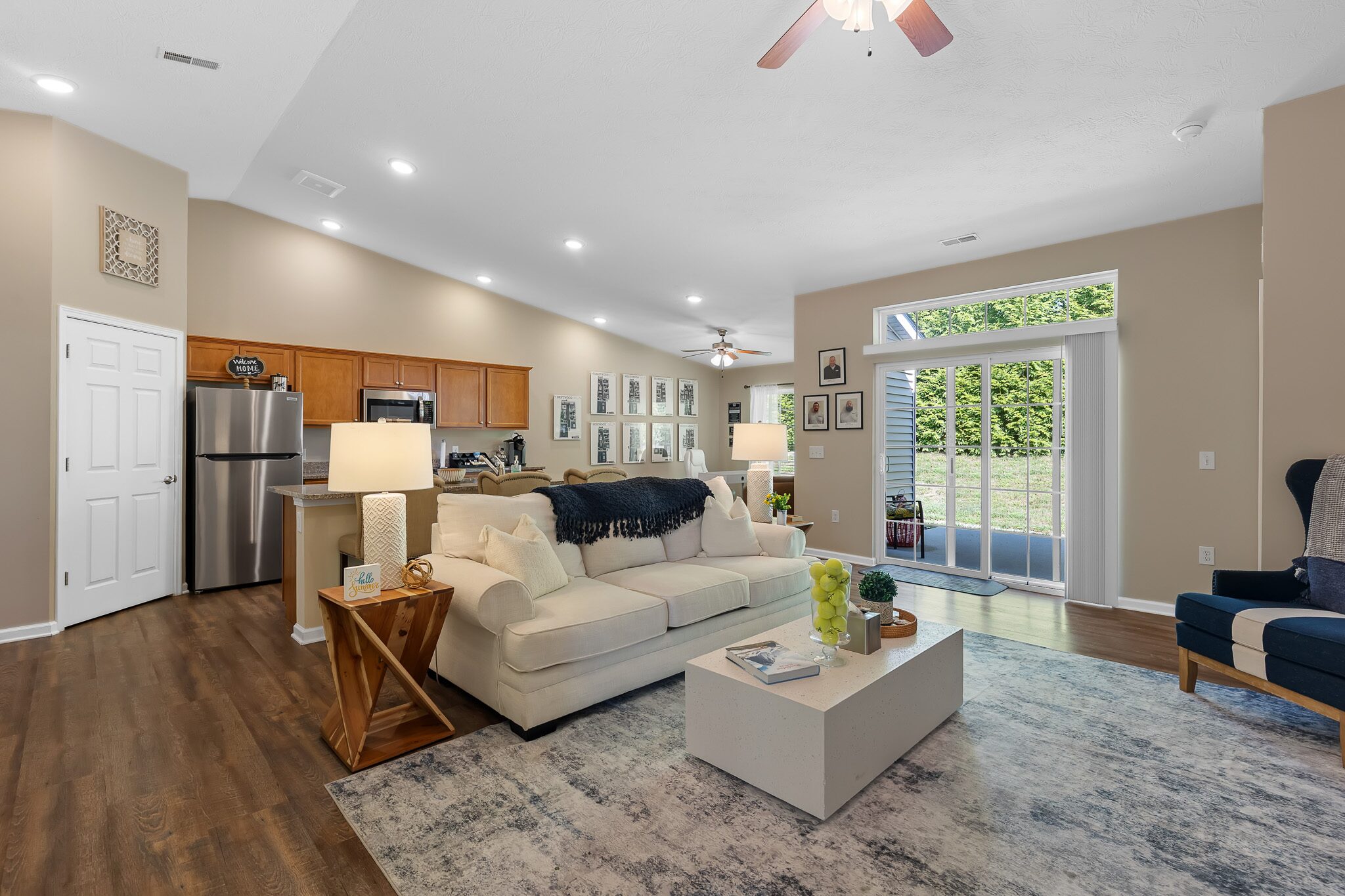 Open-concept living room and kitchen with neutral decor, light-colored sofa, modern coffee table, and sliding glass doors leading to a backyard.