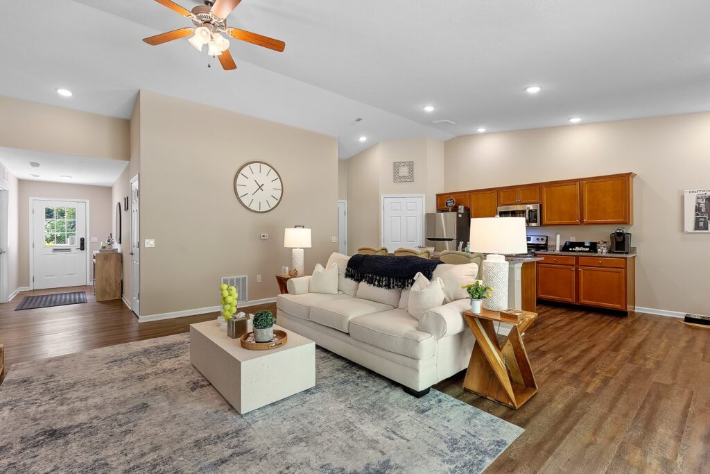Open-plan living room and kitchen with wooden floors, beige walls, a white sofa, wooden furniture, and ceiling fan with lights.