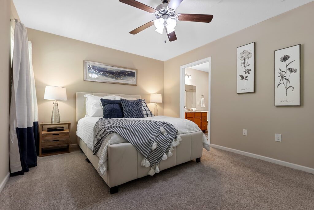 A neatly made bed with decorative pillows and throw blanket in a carpeted bedroom with neutral walls, bedside tables, lamps, and wall art; bathroom visible through open door.