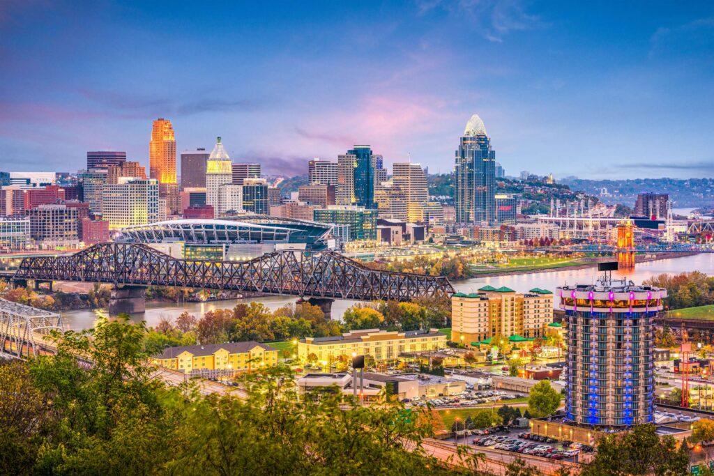 Cincinnati city skyline at dusk, with illuminated buildings, a river, bridges, and a stadium visible in the foreground.
