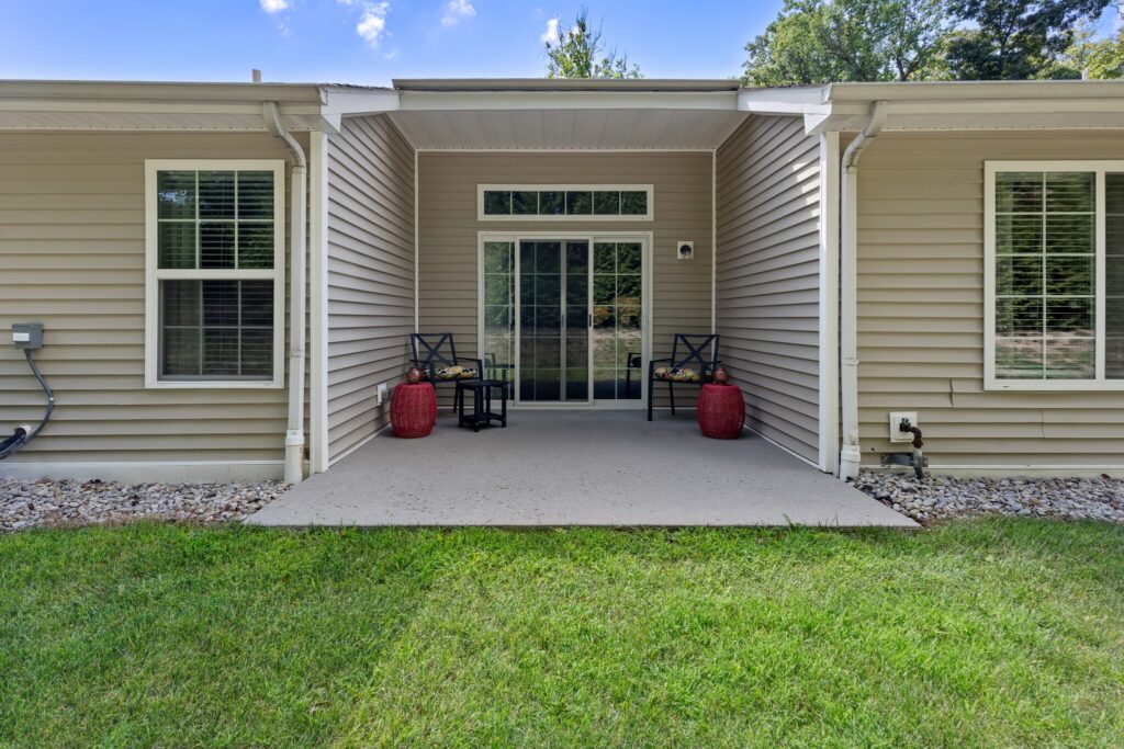 Concrete patio with two black chairs and red side tables outside a beige house with white trim and sliding glass doors, bordered by gravel and green lawn.