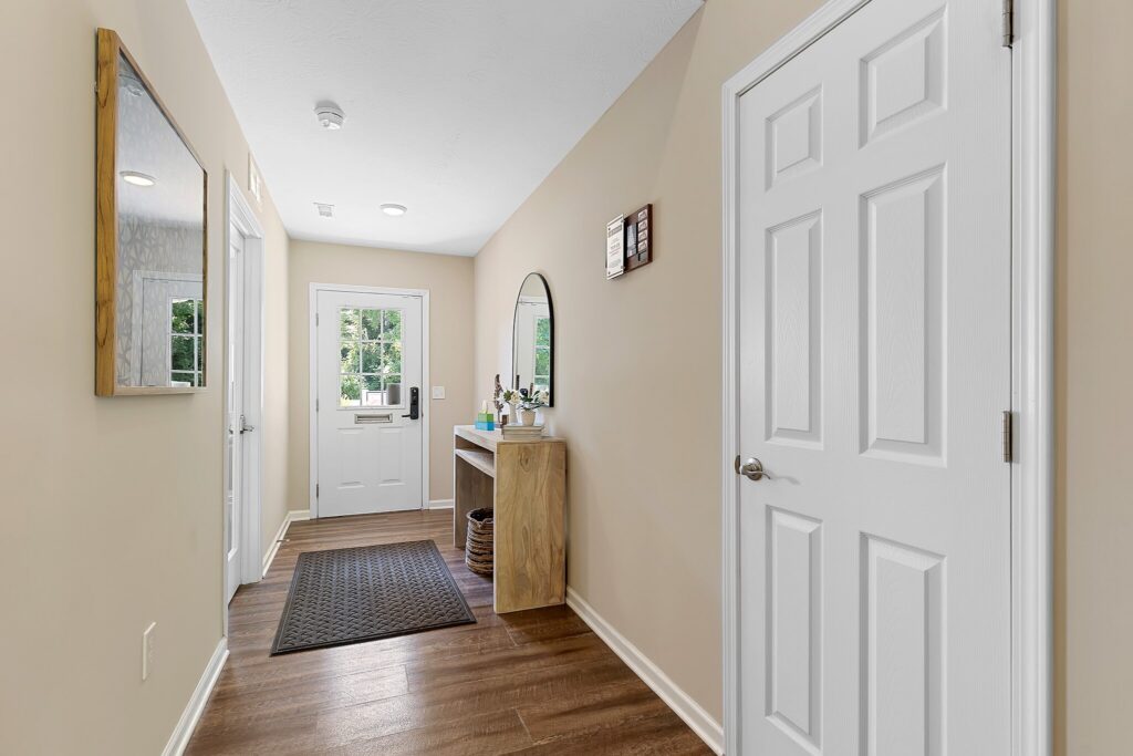A hallway with beige walls, wood flooring, a white door, a wall mirror, a small table with a round mirror, and a black mat near the entrance.