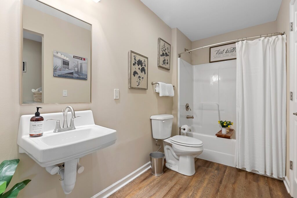 A clean bathroom with a sink, toilet, and shower-tub combo; neutral walls, wood-look flooring, and simple decor including framed art and a small plant on the tub tray.