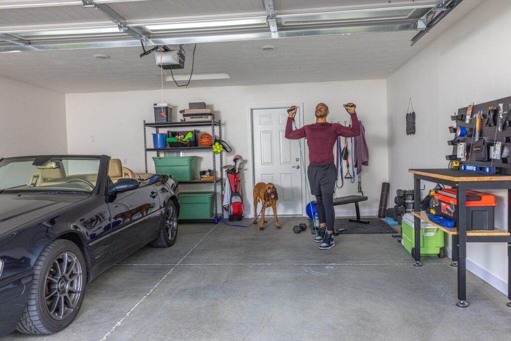 A man does pull-ups on a bar in a clean garage with a dog nearby, a convertible car, tools, and sports equipment visible.