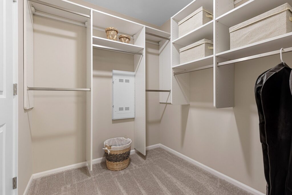Walk-in closet with white shelving, hanging rods, storage baskets, a laundry hamper, and an electrical panel on the back wall. Carpeted floor and beige walls.